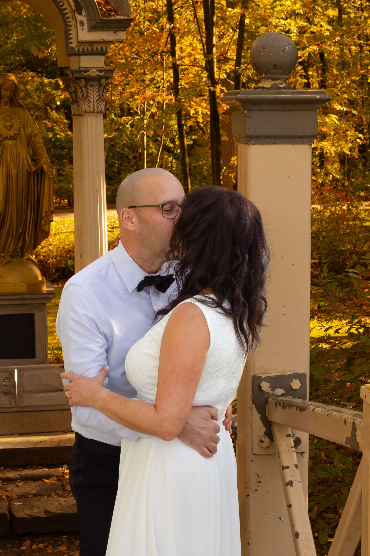 Bride and groom sharing a kiss on a bridge with golden autumn foliage and statue in background.