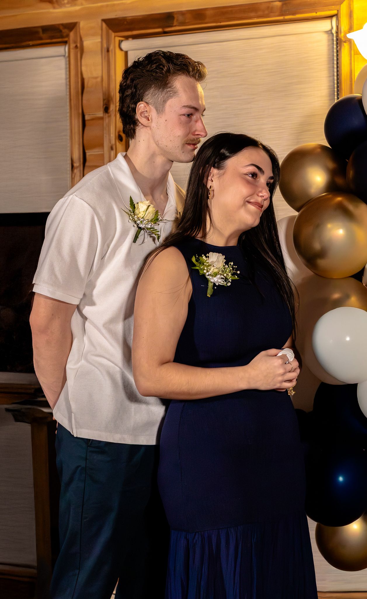 Young couple in navy and white formal attire with boutonnieres pose together at balloon-decorated wedding reception.