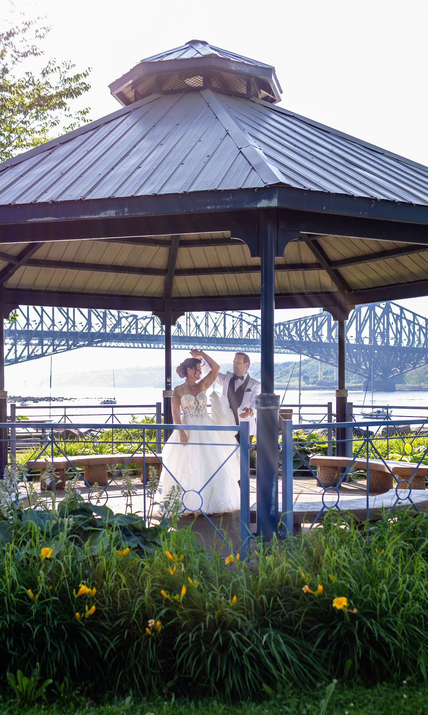 Bride and groom dancing in a gazebo with a scenic river bridge backdrop, surrounded by lush greenery and yellow flowers.