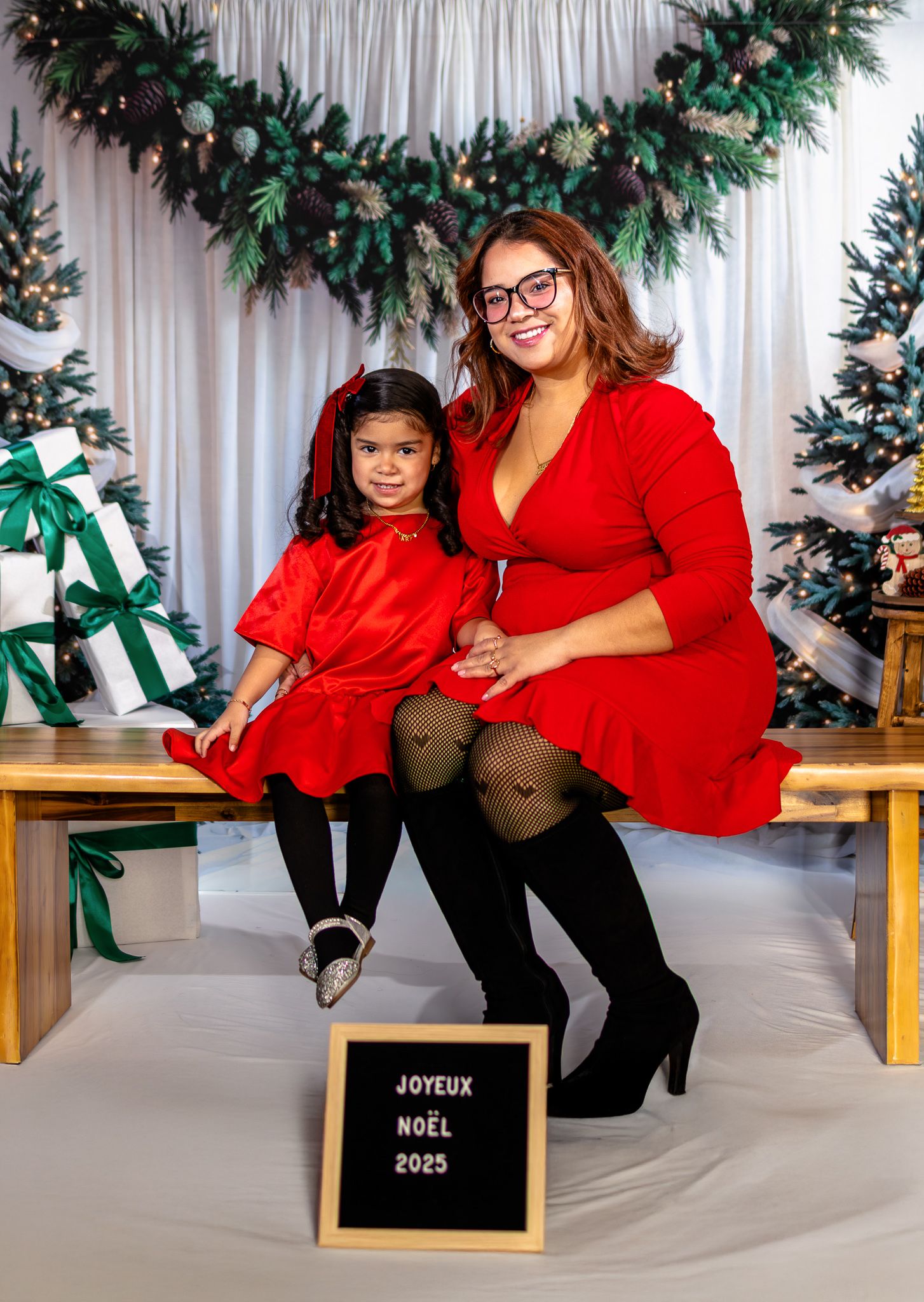 Mother and daughter in matching red dresses posing with Joyeux Noel sign in festive Christmas photo session.