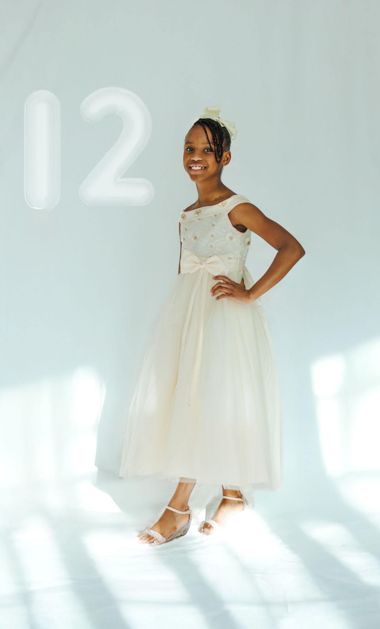 Happy young girl in elegant white tulle dress posing beside number 12 balloon on light background.