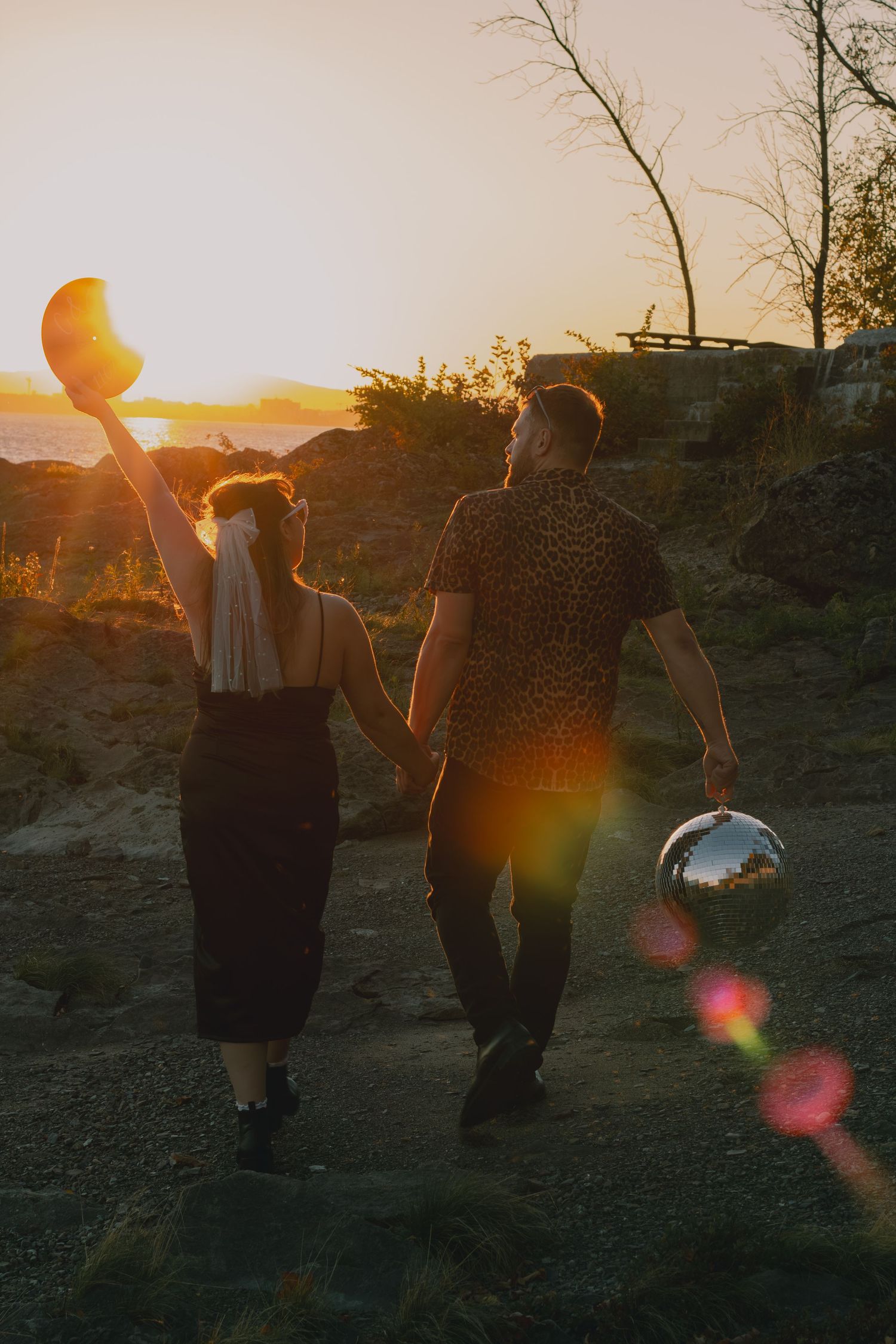 Couple holding hands walking toward sunset on rocky terrain, woman raising hat, man carrying disco ball, warm golden light.