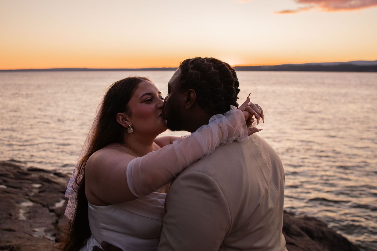 Couple sharing a romantic kiss at sunset by the ocean, woman in white dress embracing man on rocky shoreline.