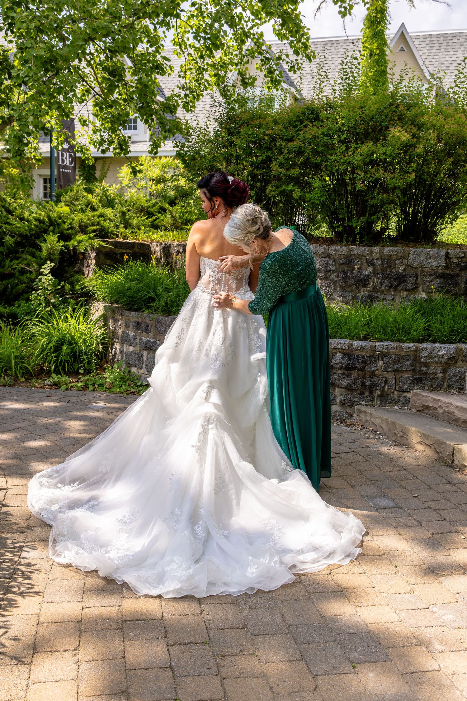 Bride in white ball gown getting dressed by woman in green dress in a lush garden setting.