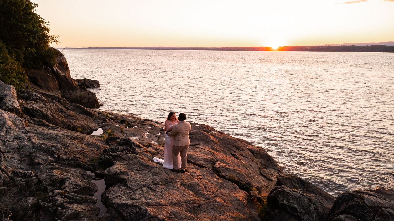 Couple embracing on rocky shoreline watching golden sunset over calm lake waters.