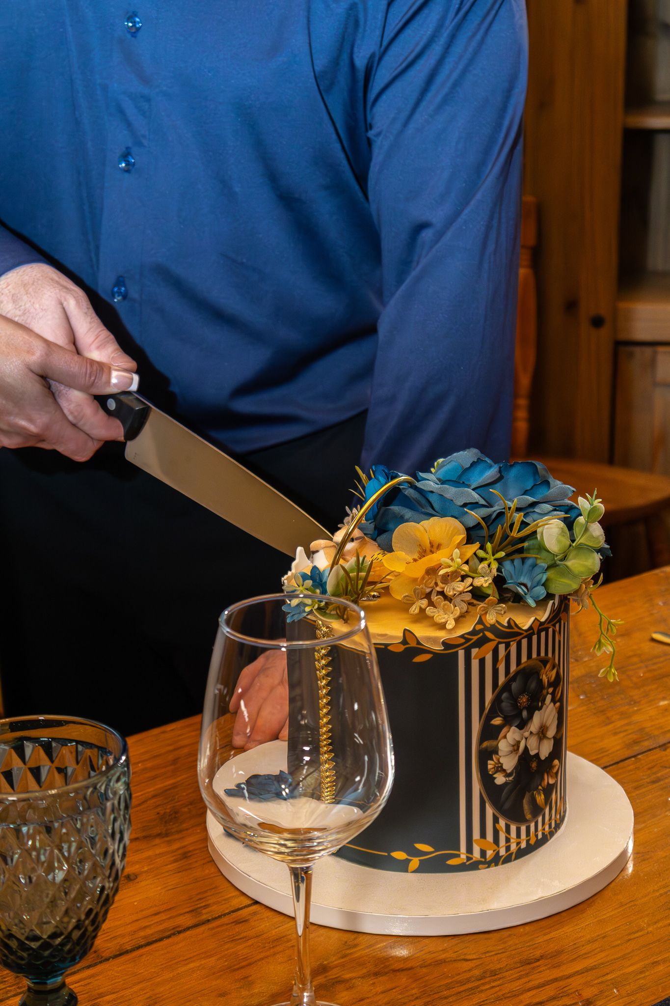 Man in blue shirt cutting elegant black and gold floral cake beside wine glass on wooden table.