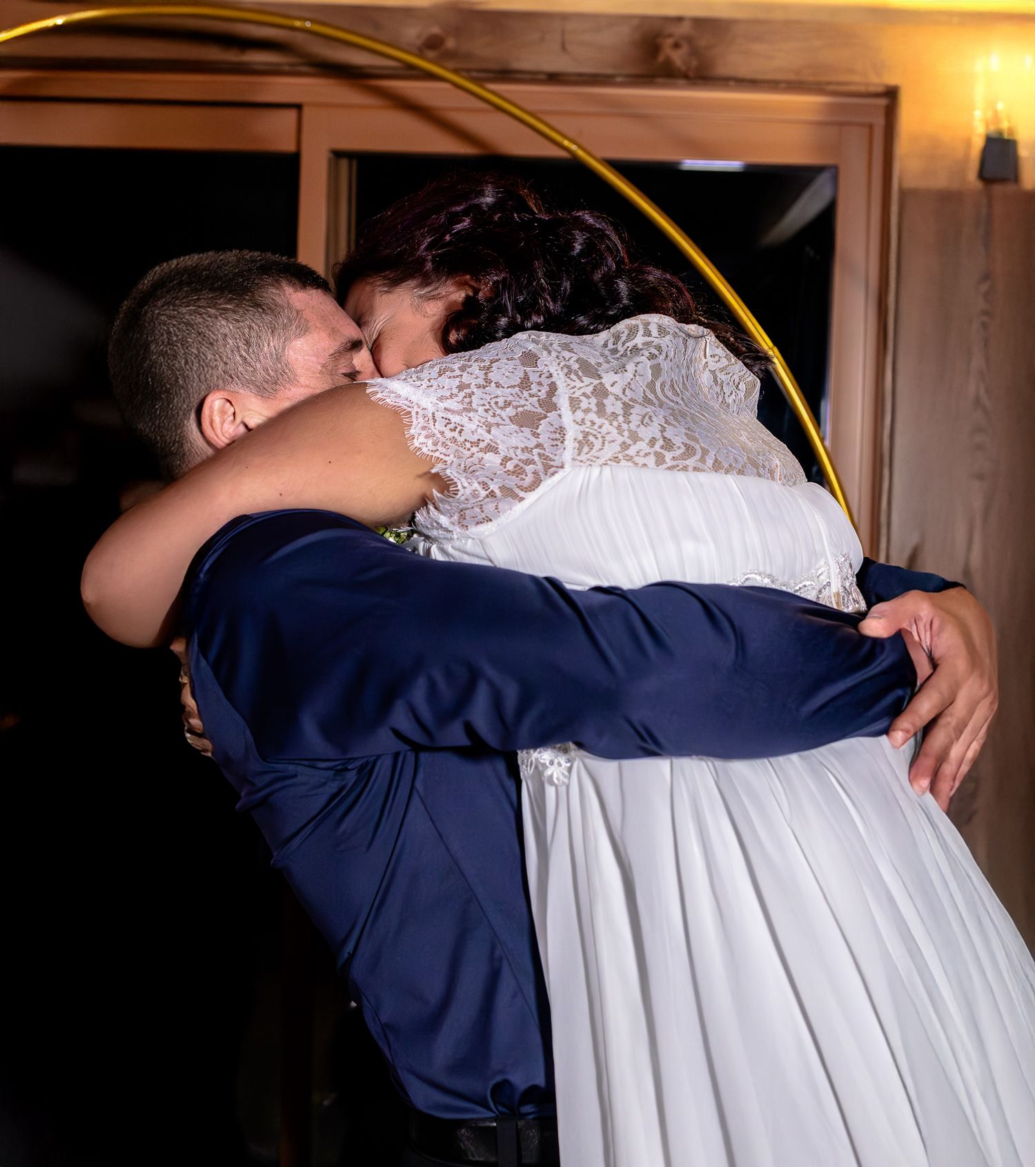 Bride and groom share passionate kiss and embrace under golden hoop arch at wedding celebration.
