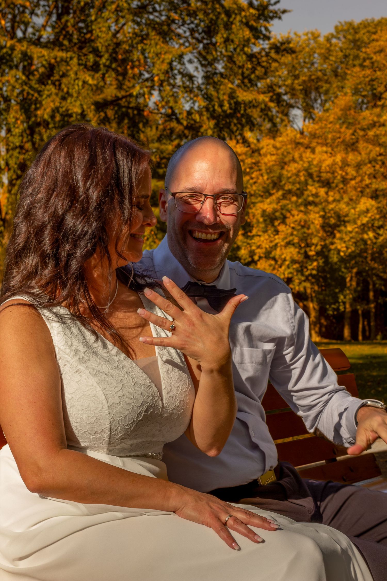Laughing groom with glasses sits on a bench as bride shows off her wedding ring in autumn park.