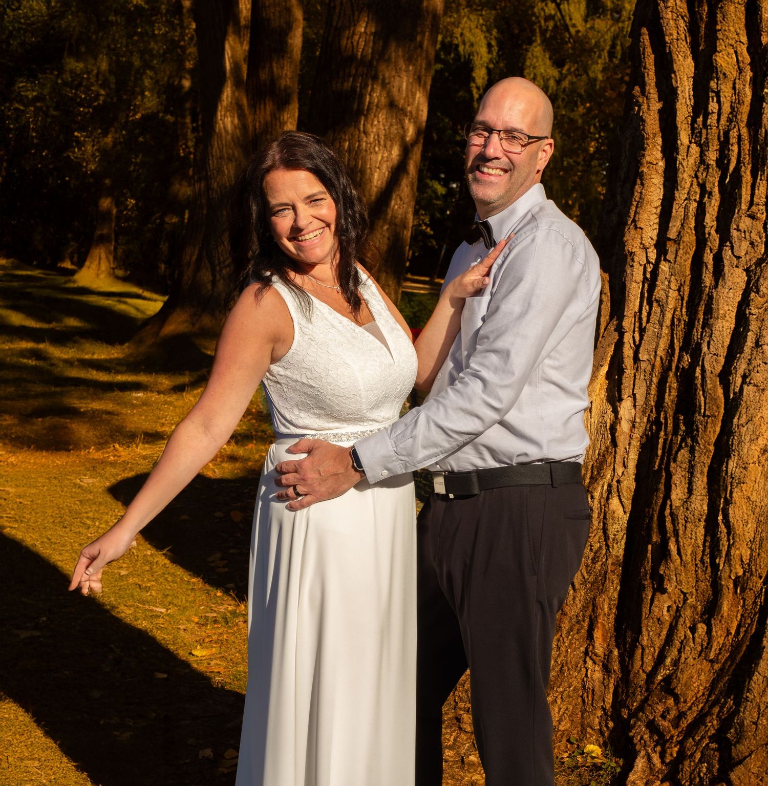 Smiling wedding couple posing against a large tree trunk in warm golden autumn sunlight.