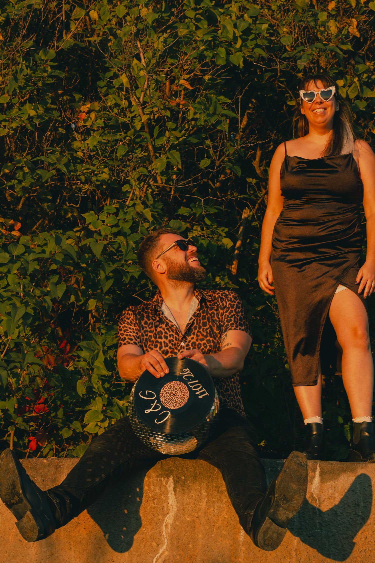 Couple poses outdoors at golden hour, man seated with drum, woman standing in black dress.
