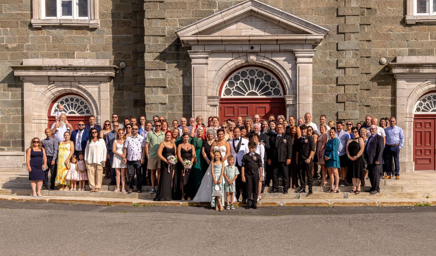 Large wedding group photo outside historic stone church with bride and groom centered among guests on sunny day.