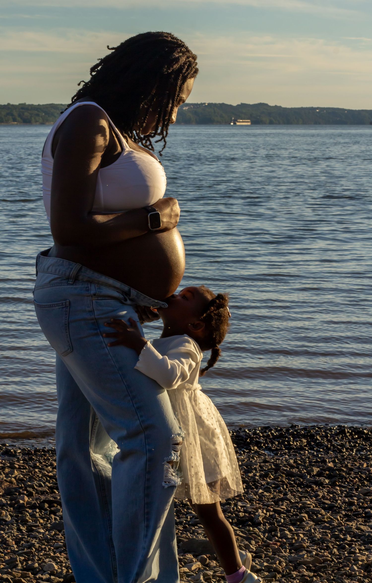Pregnant Black woman in white top and jeans stands on a pebble beach as a young girl kisses her baby bump.