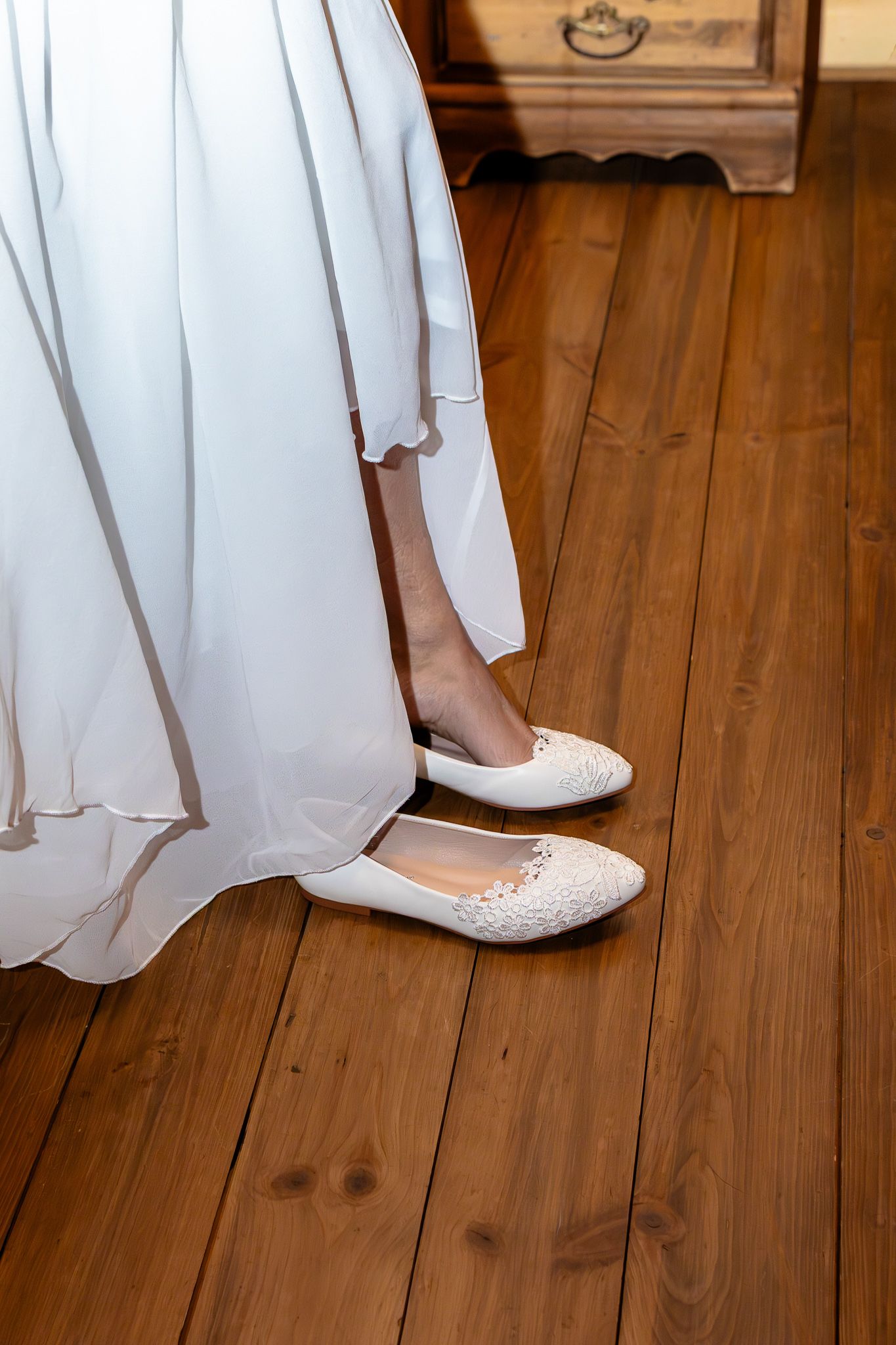 Close-up of a bride's white lace flat shoes peeking beneath her flowing white wedding dress on hardwood floor.