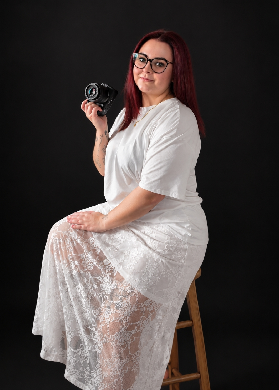 Woman with red hair and glasses in white lace dress sits on stool holding a camera against black background.