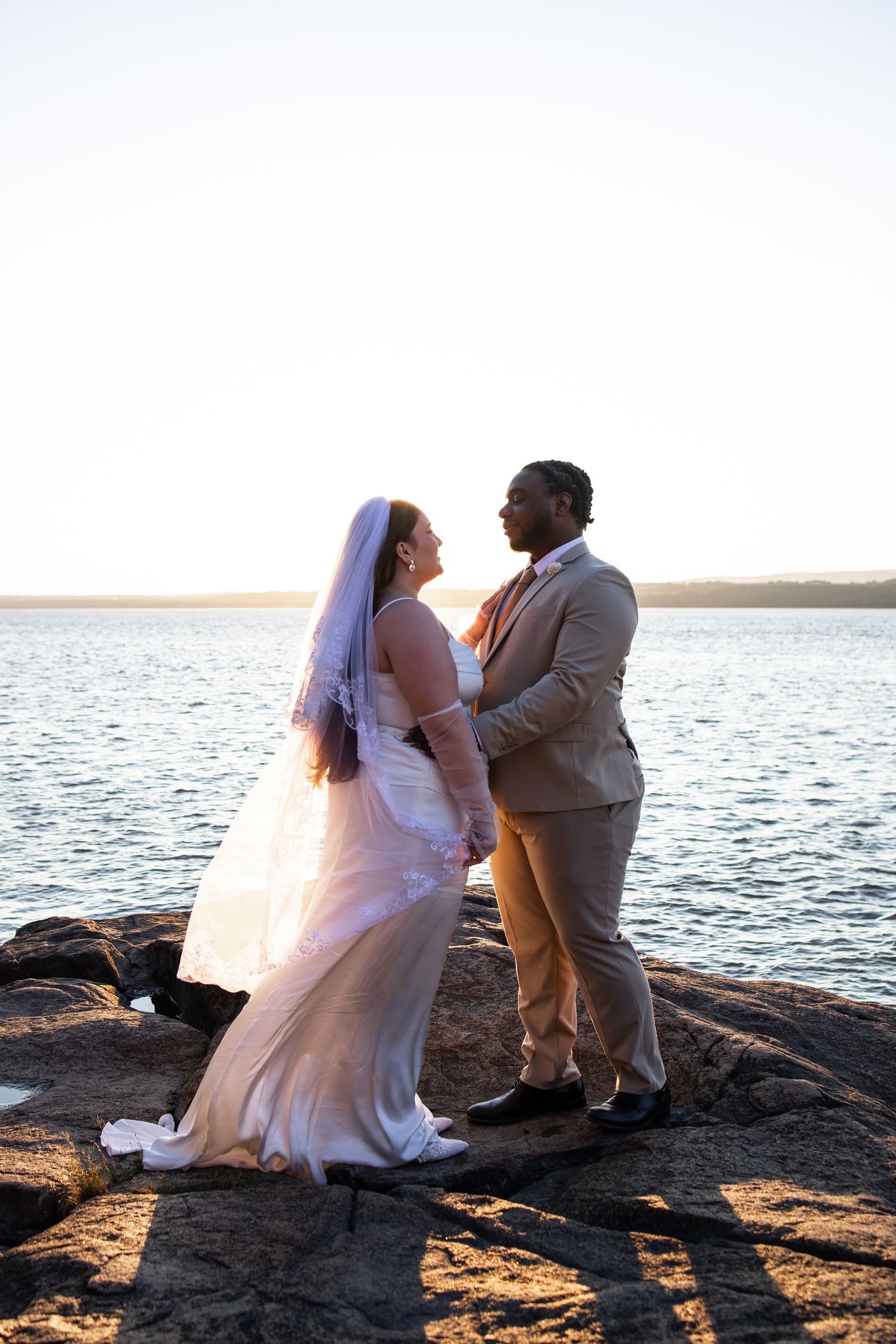Interracial couple in wedding attire sharing a romantic moment on rocky shoreline at sunset.