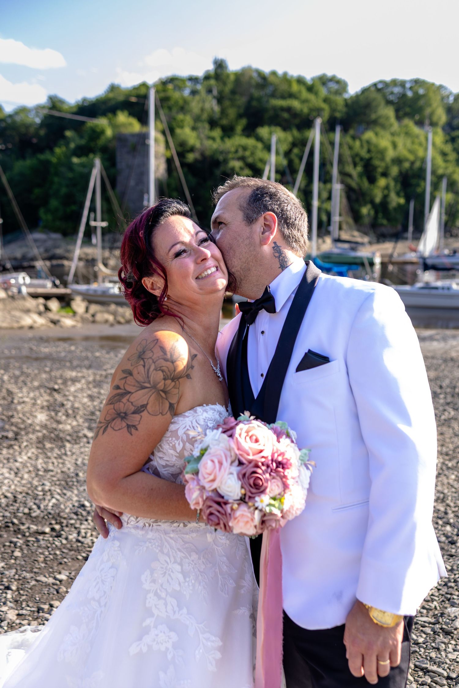 Bride and groom share a romantic kiss at a waterfront marina, bride holding pink bouquet in white lace gown.