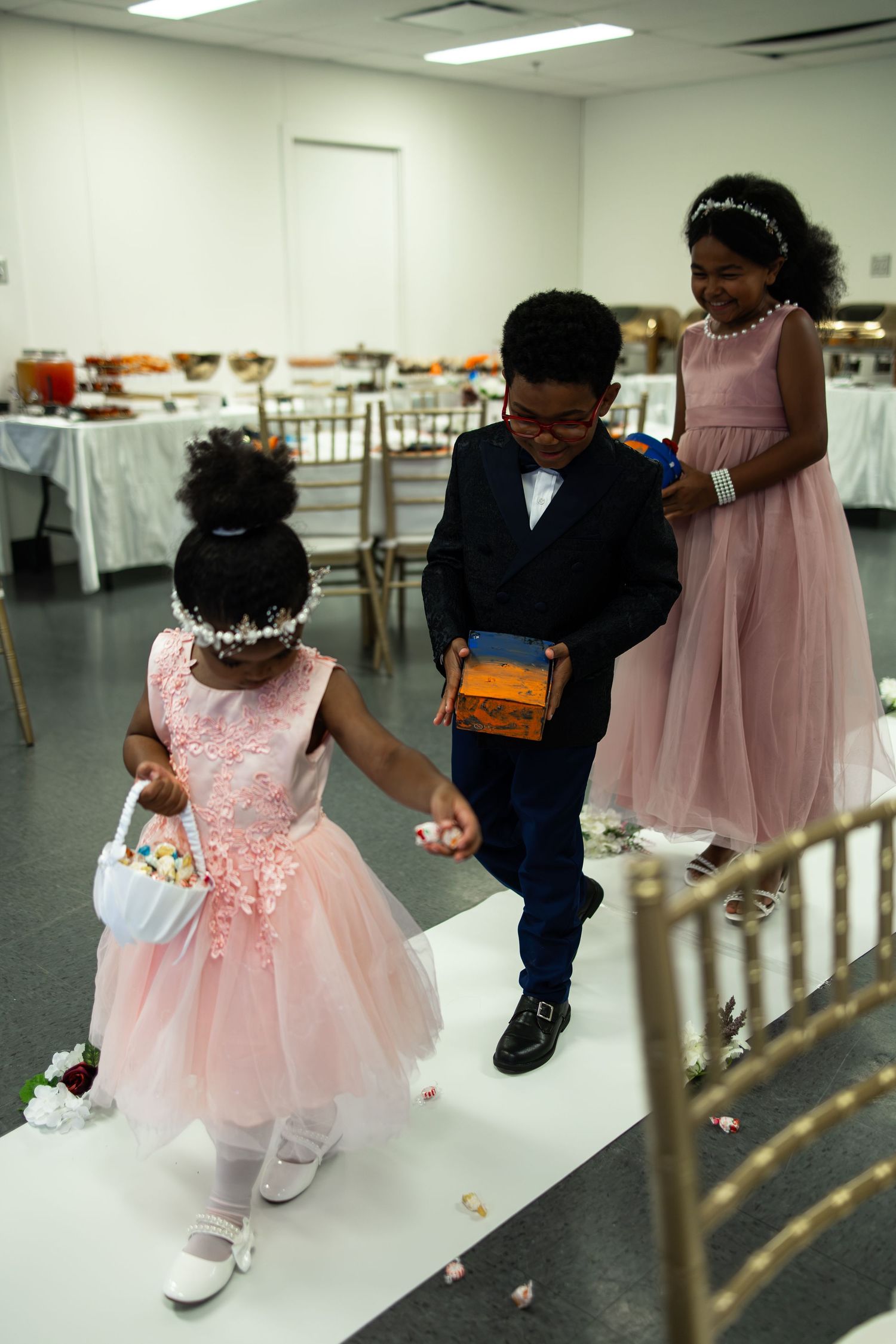 Three children in formal attire scatter flower petals at a wedding reception, with a boy holding a gift box.