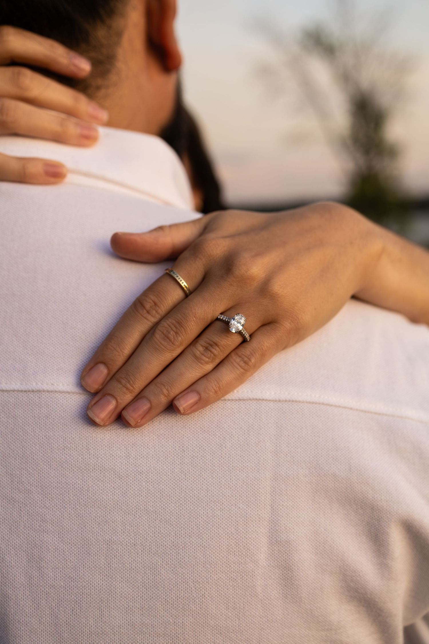 Close-up of a woman's hand with a diamond engagement ring and gold band resting on a man's shoulder.
