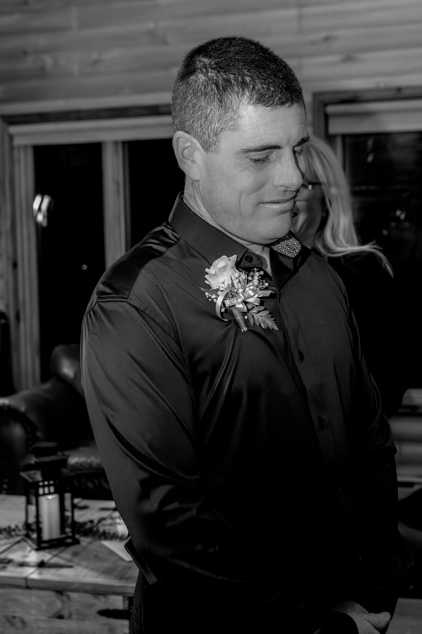 Black and white portrait of groom in dark shirt with floral boutonniere in rustic wood venue.