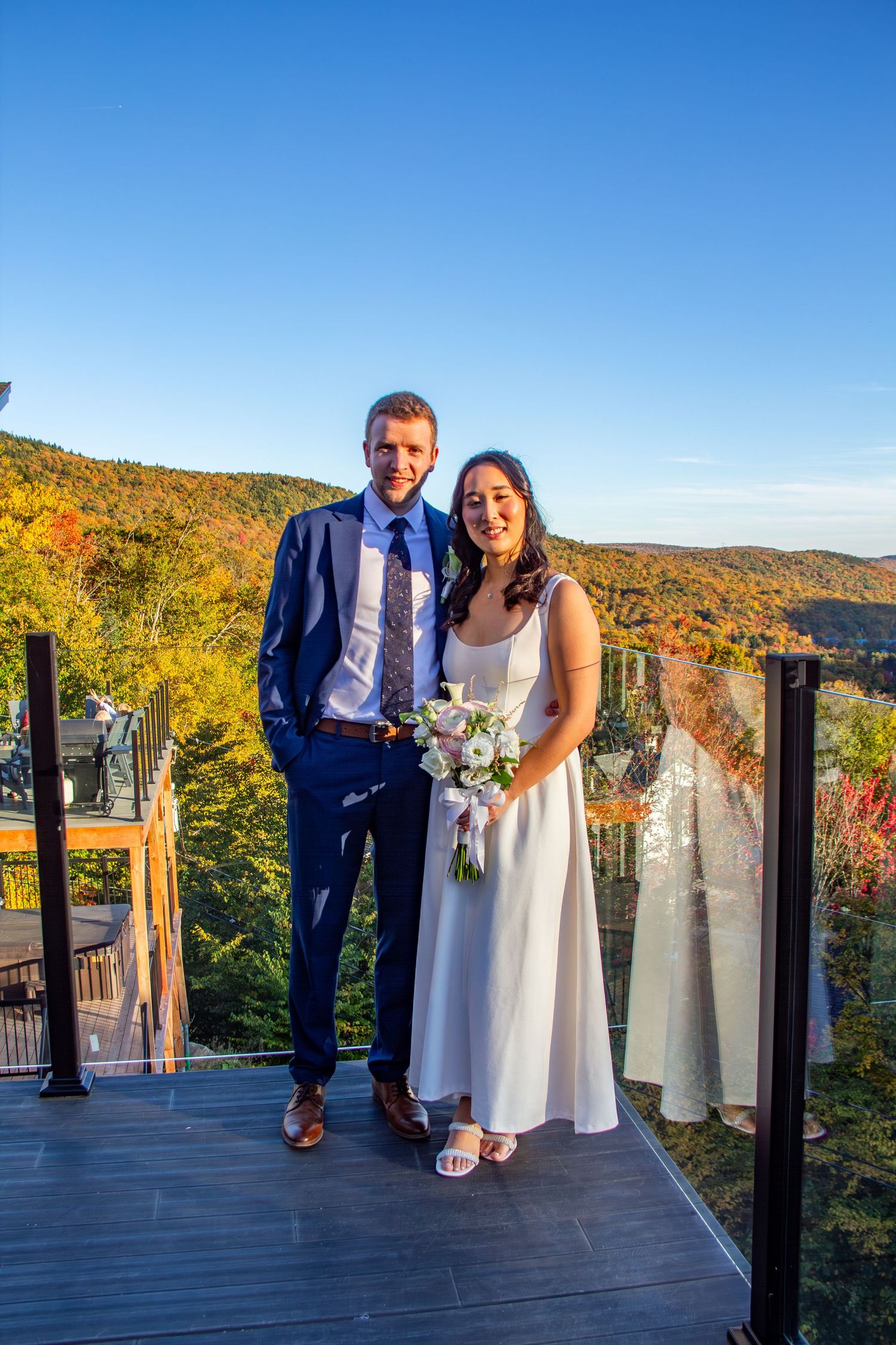 Smiling bride and groom posing on a deck with colorful autumn mountain foliage backdrop.