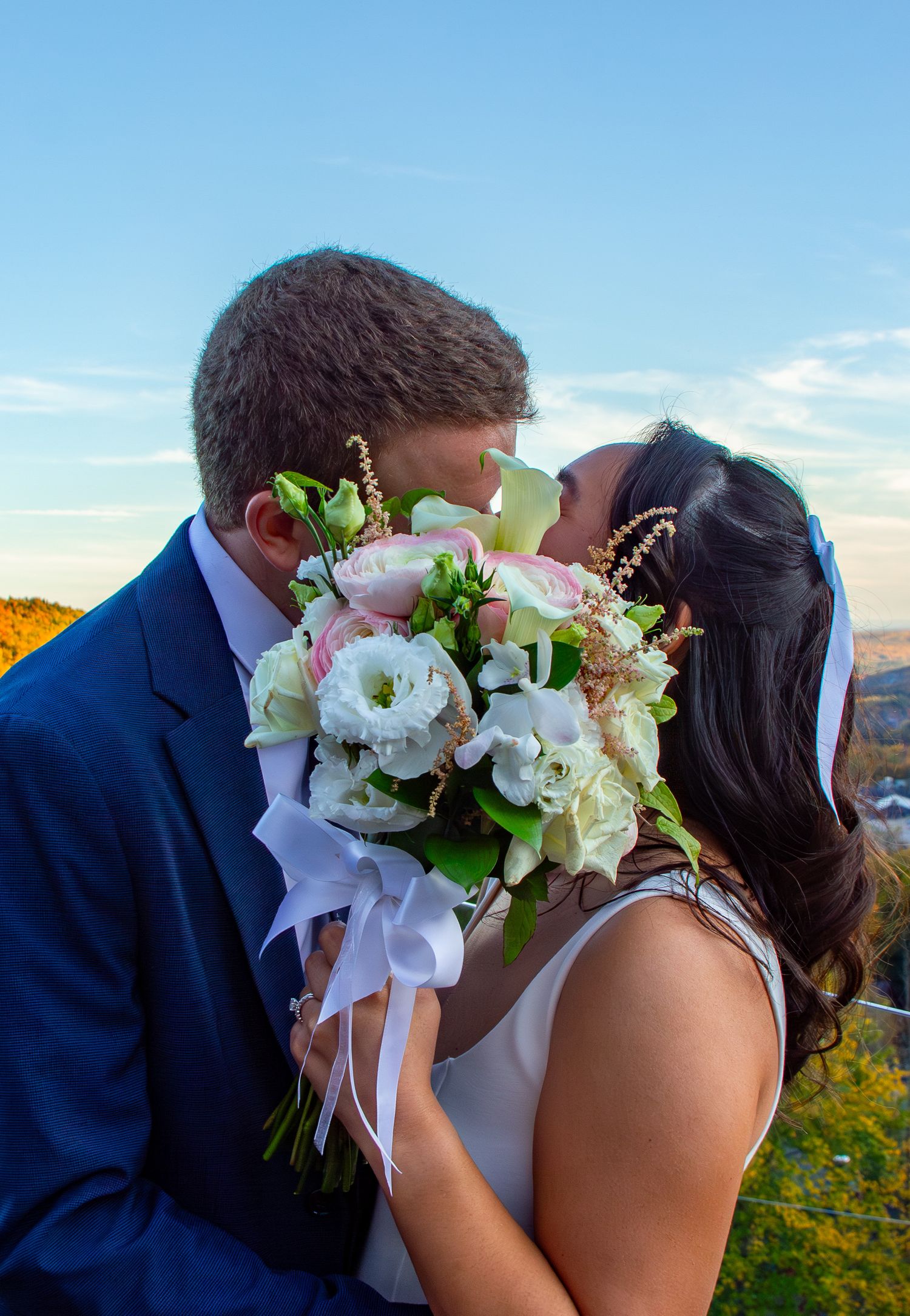 Bride and groom share a kiss hidden behind a beautiful white and pink wedding bouquet at sunset.