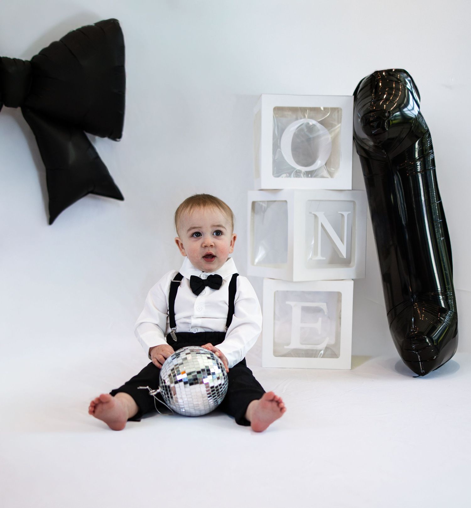 Baby boy in suspenders and bow tie holds disco ball near letter blocks spelling ONE for his first birthday photo.