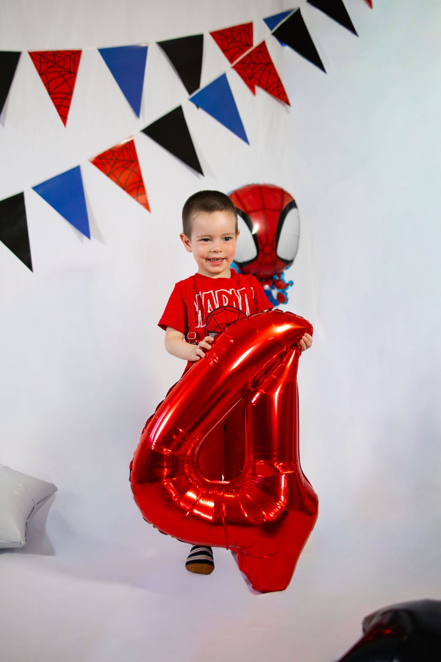 A young boy in a red shirt holds a large red number 4 balloon at a Spiderman-themed birthday party.