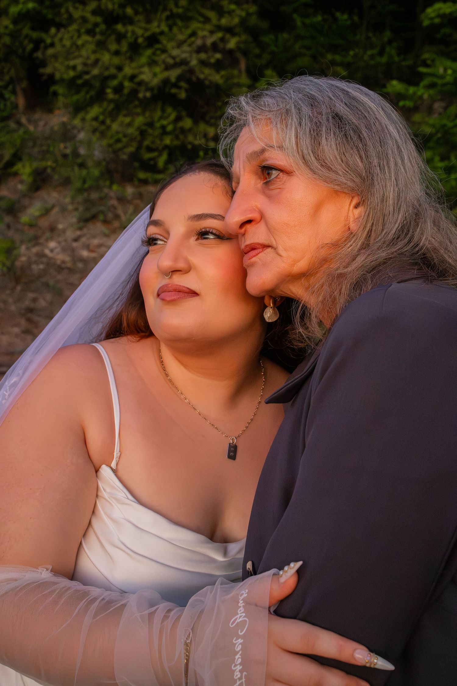 A bride in a white dress and veil shares a tender moment with an older woman who kisses her cheek at golden hour.