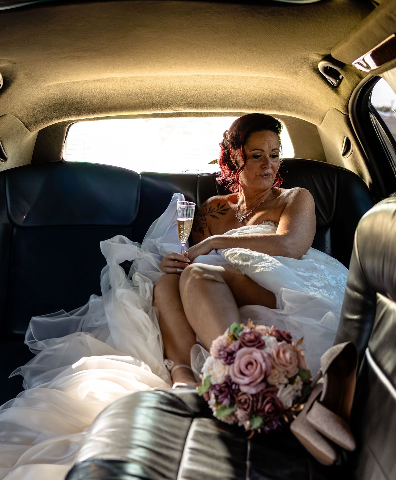 Bride in white wedding dress sitting in limousine reading a card, holding champagne, with pink floral bouquet nearby.