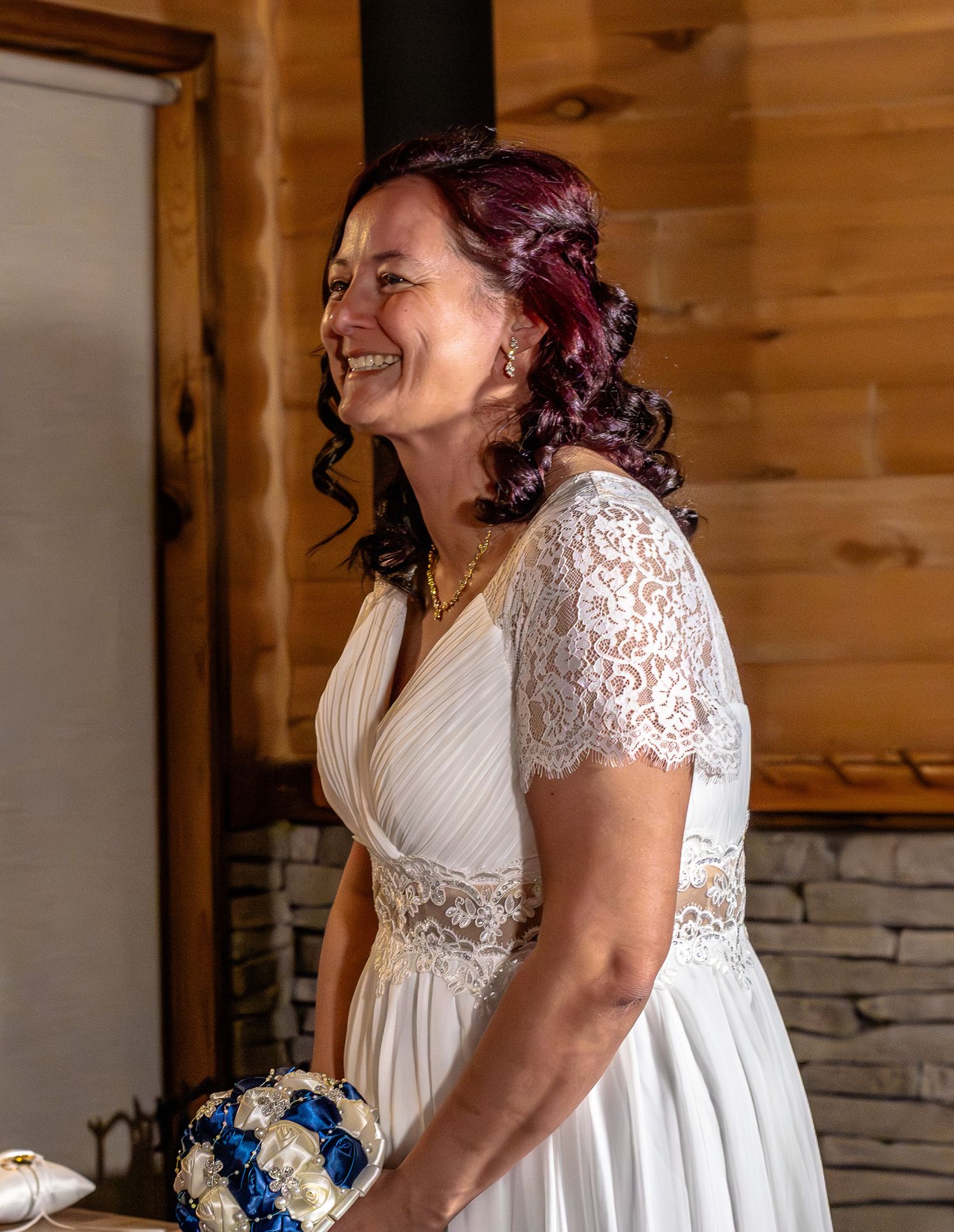 Radiant bride with red hair laughs joyfully in white lace wedding gown holding blue and white bouquet.