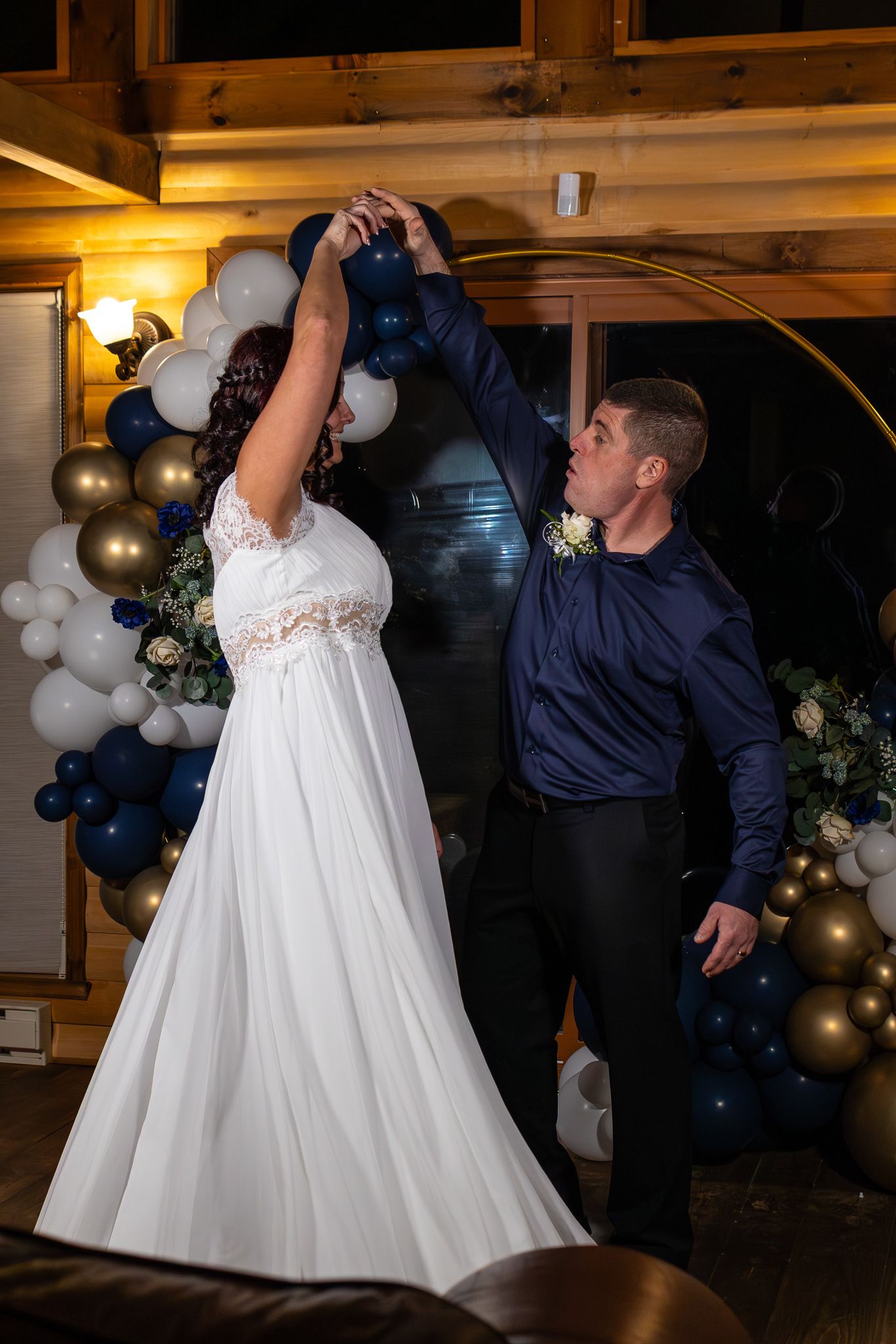 Groom spins bride in white lace gown under a gold circular arch decorated with navy and gold balloons.