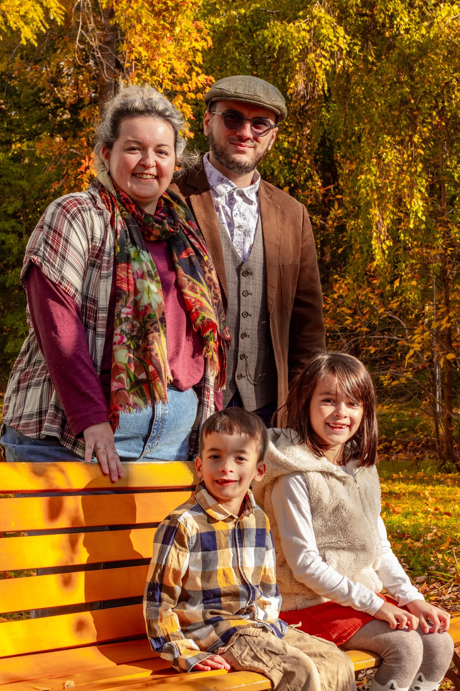 Family of four posing on yellow bench surrounded by vibrant autumn foliage in a sunny park.