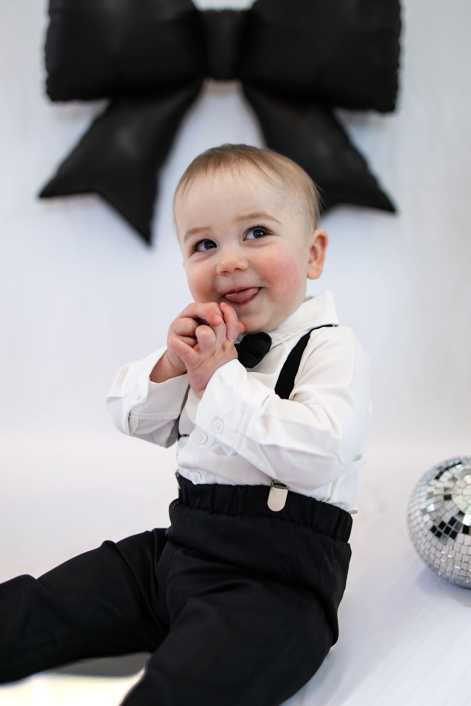 Smiling baby boy in white shirt, bow tie, suspenders and black pants sits on white floor with disco ball.