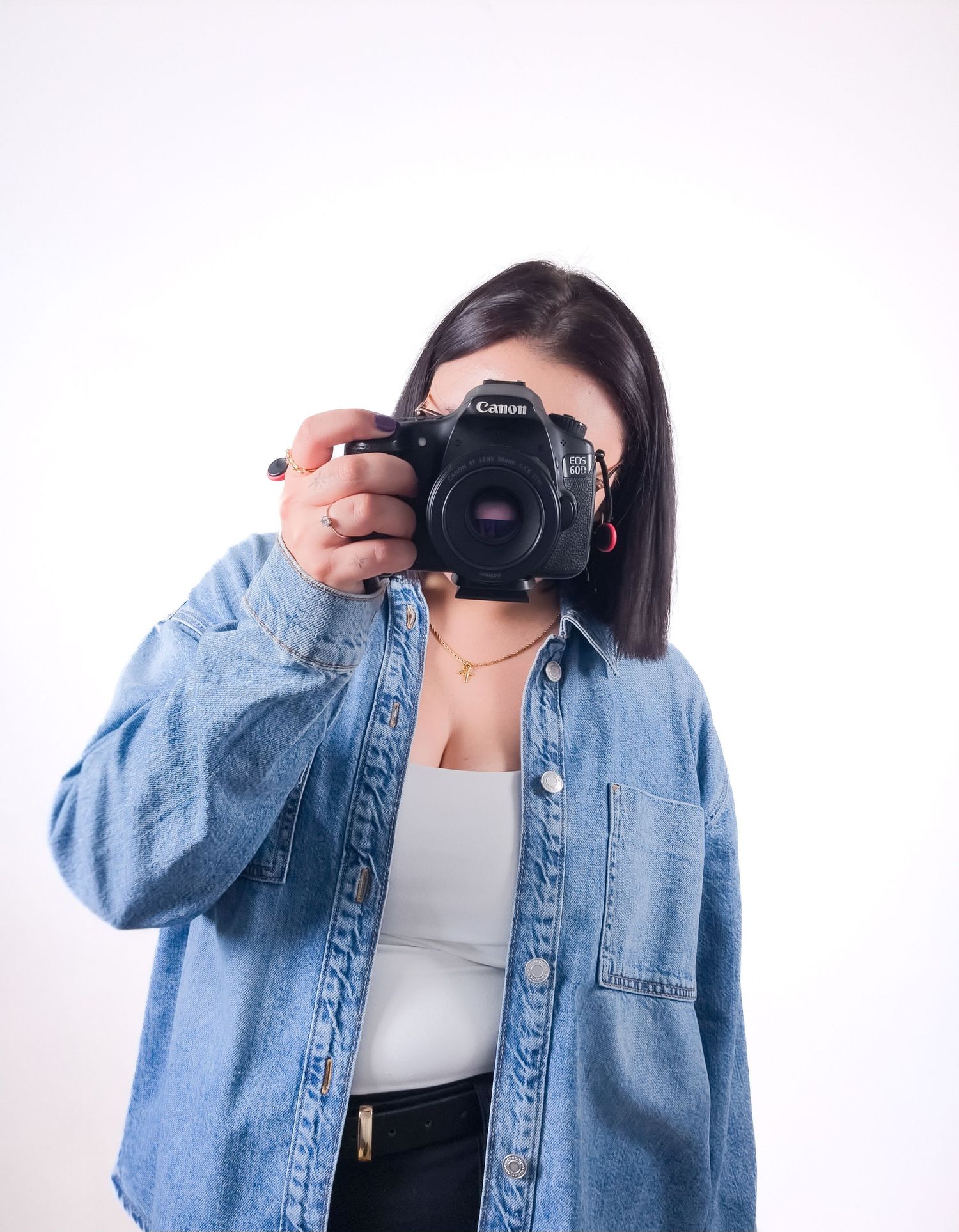 Female photographer in denim jacket aiming a Canon DSLR camera toward the viewer on white background.