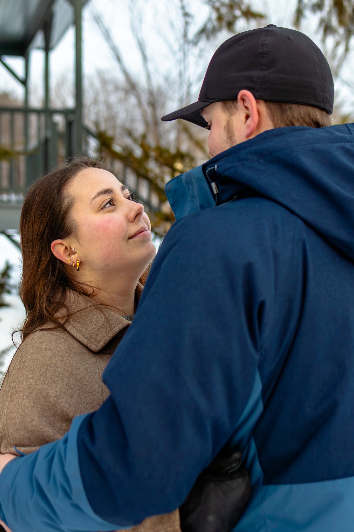 Woman gazing up lovingly at man in black cap and blue jacket while embracing outdoors in winter snowy setting.