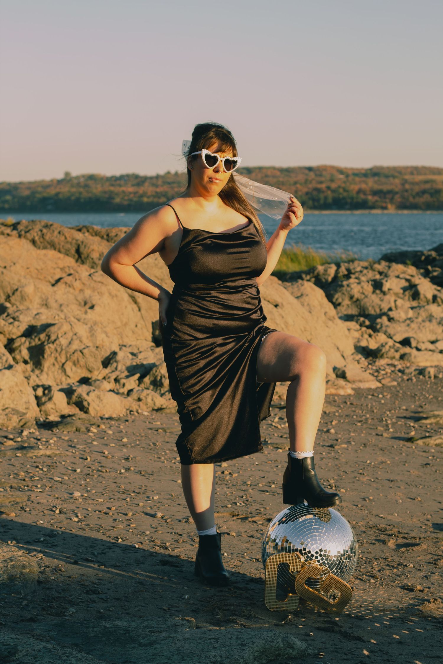 Woman in black satin dress, heart sunglasses, poses with disco ball on rocky beach at sunset.