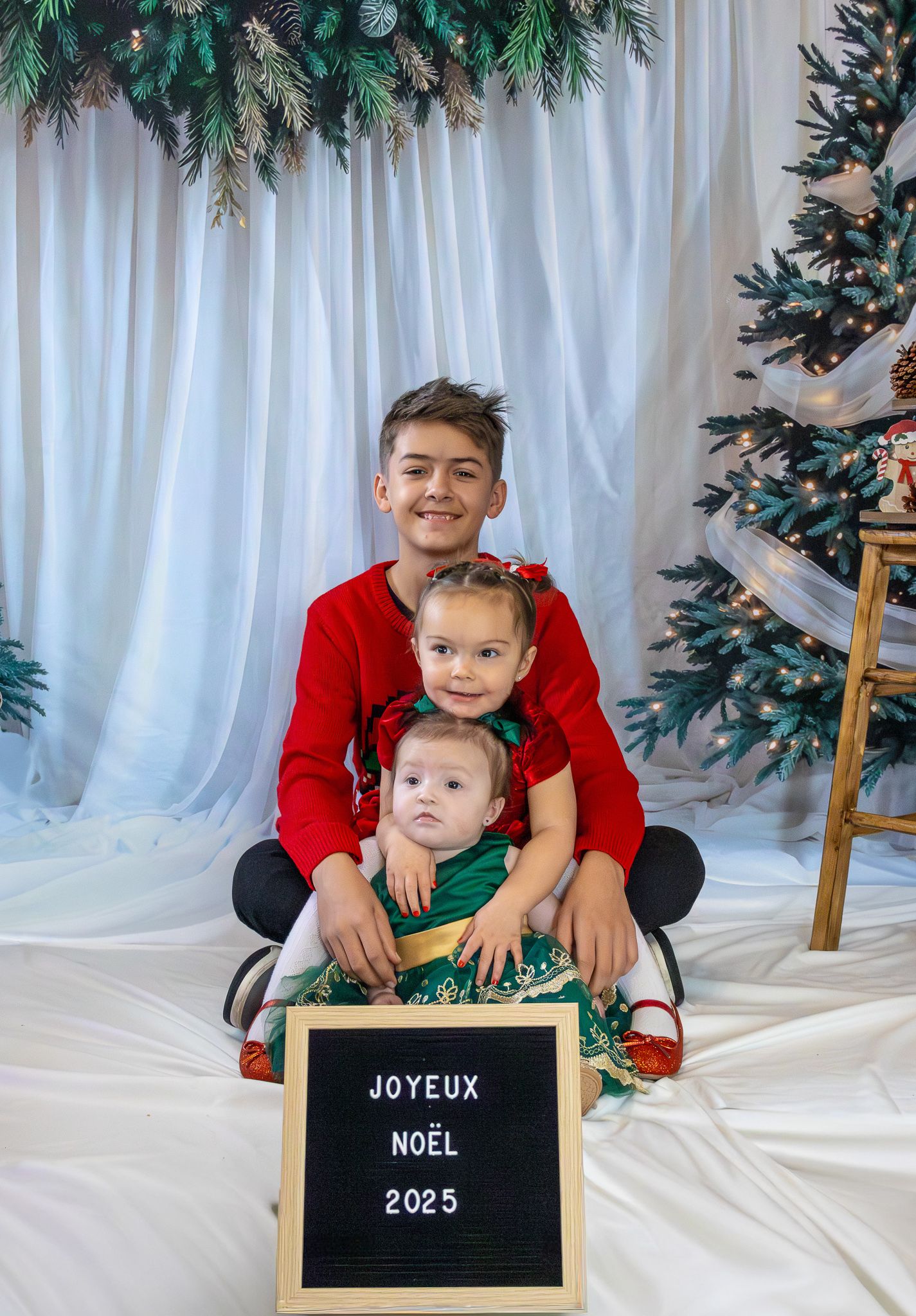 Three siblings posing together with a Joyeux Noel 2025 sign in front of a Christmas tree.