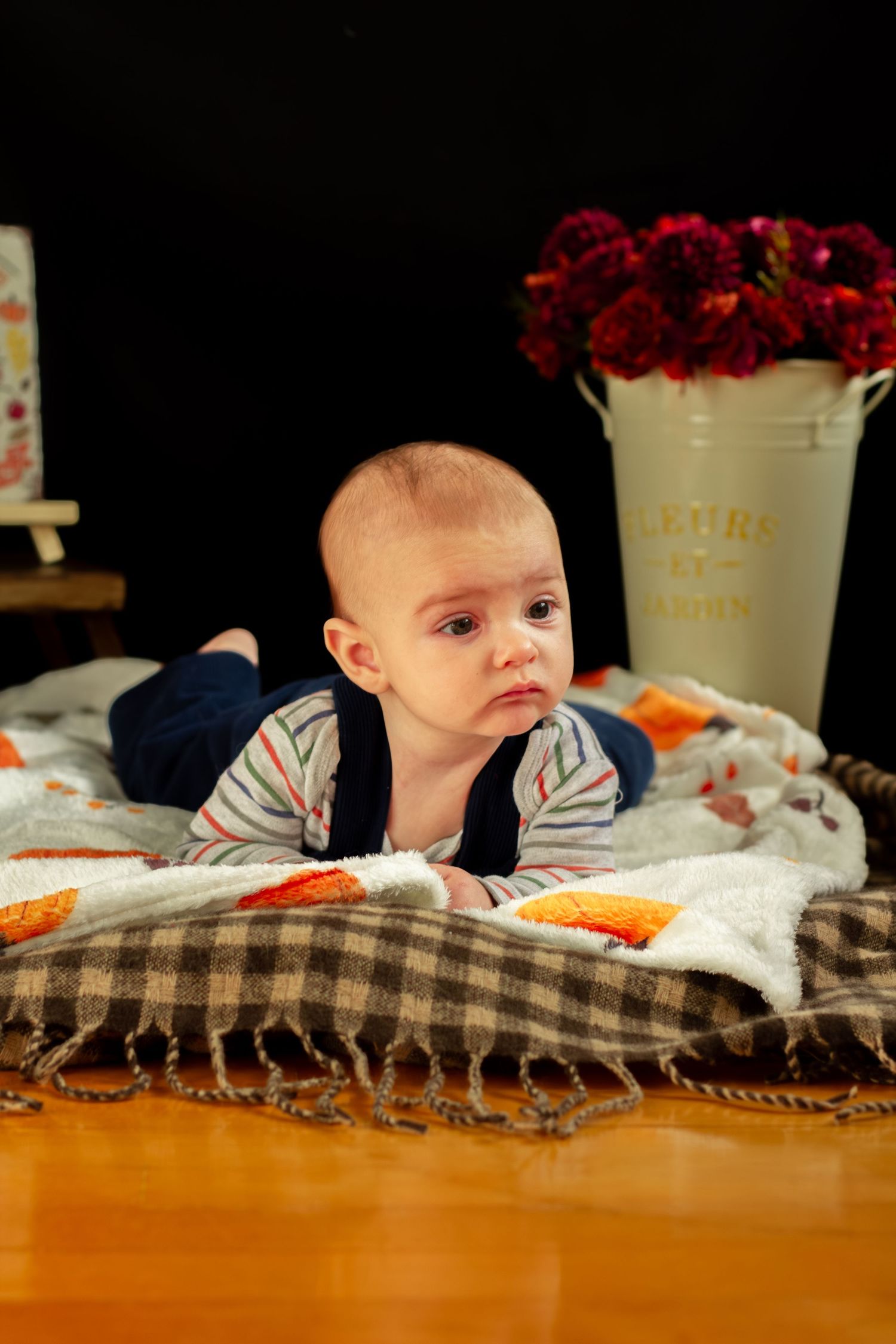 Baby in blue overalls doing tummy time on a plaid blanket with fall floral decorations.