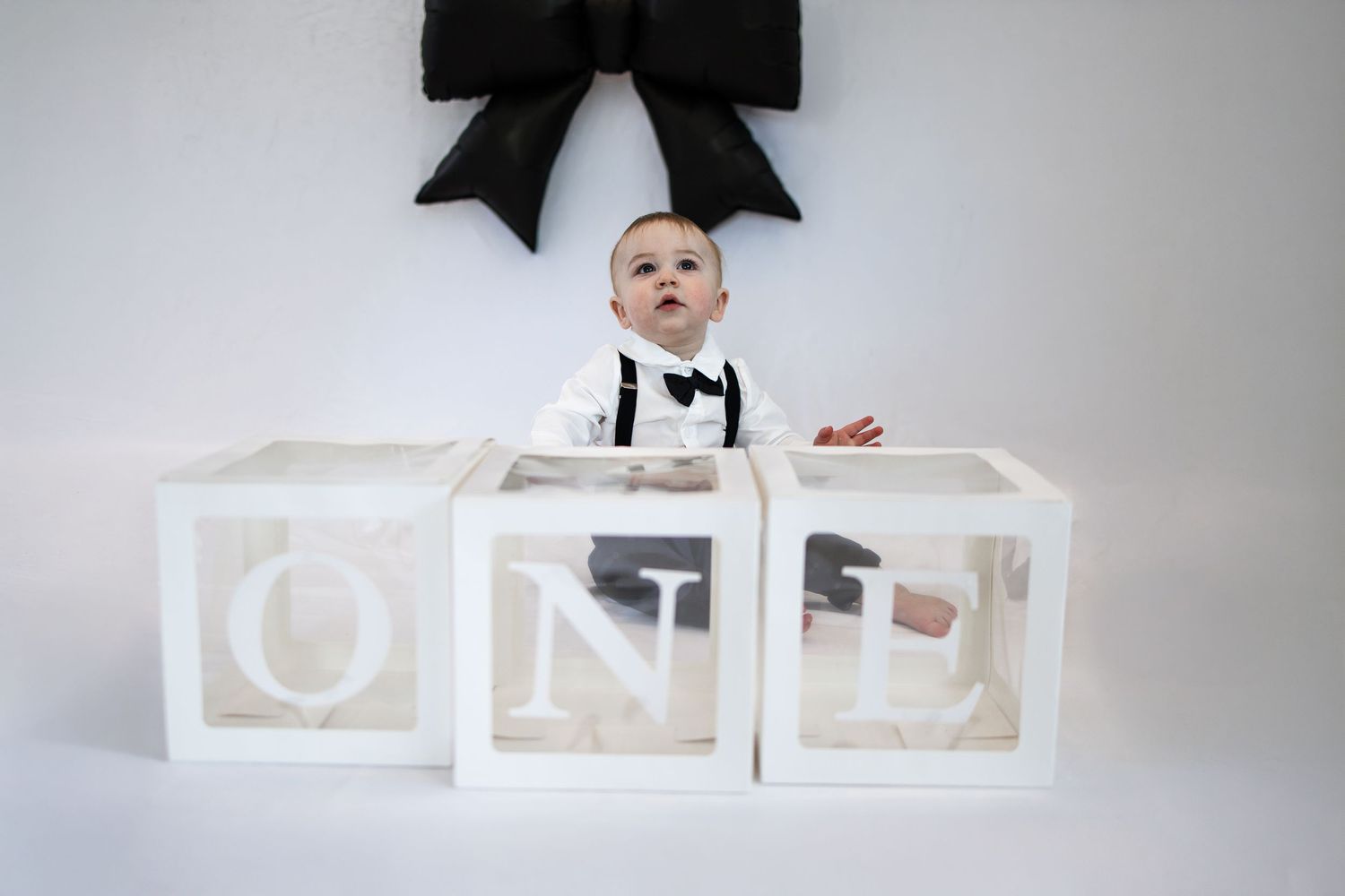 Baby in white tuxedo sits between three clear letter blocks spelling ONE, with black bow decor, for first birthday photo.