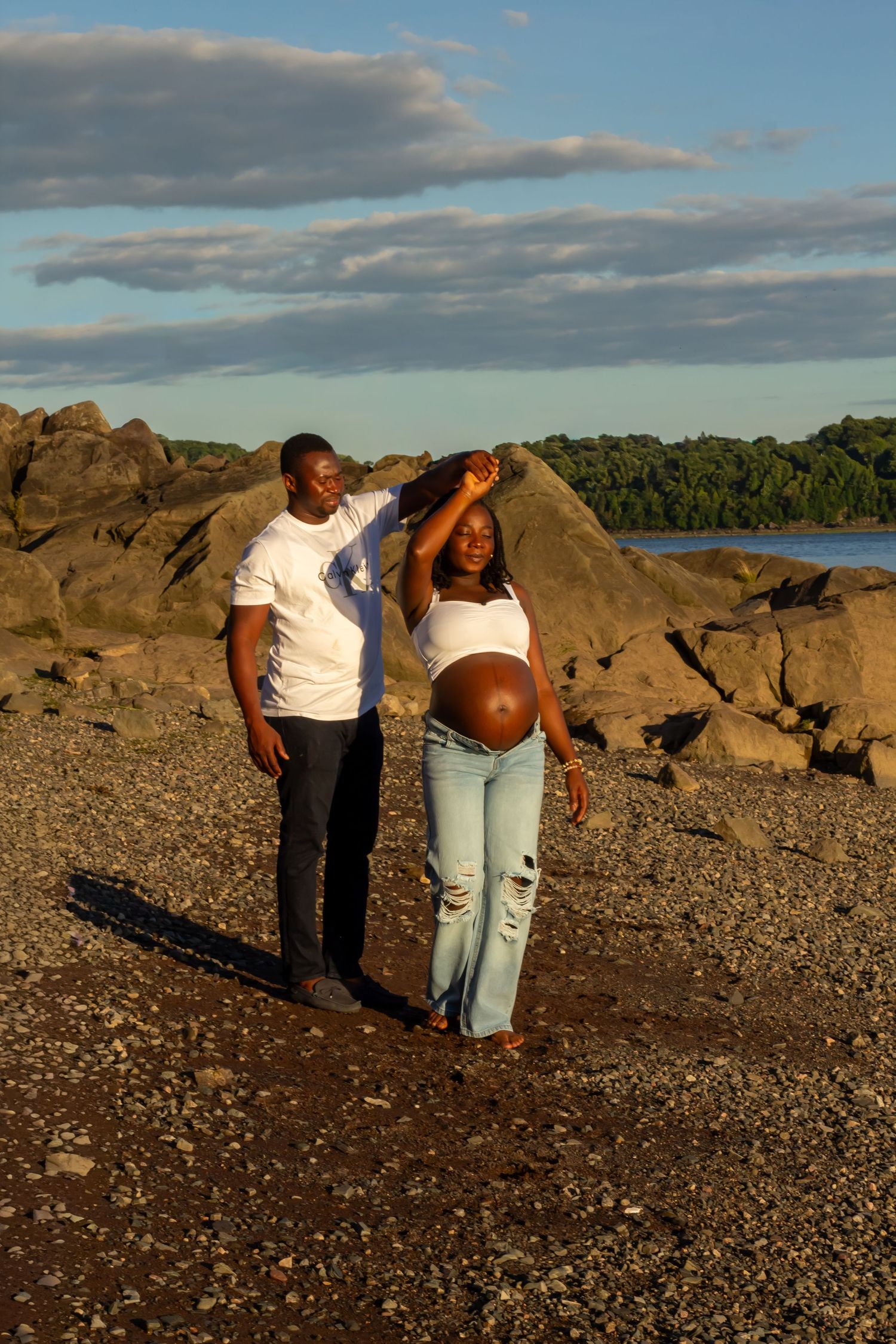 A man spins a pregnant woman in a white crop top and jeans on a rocky beach at sunset.