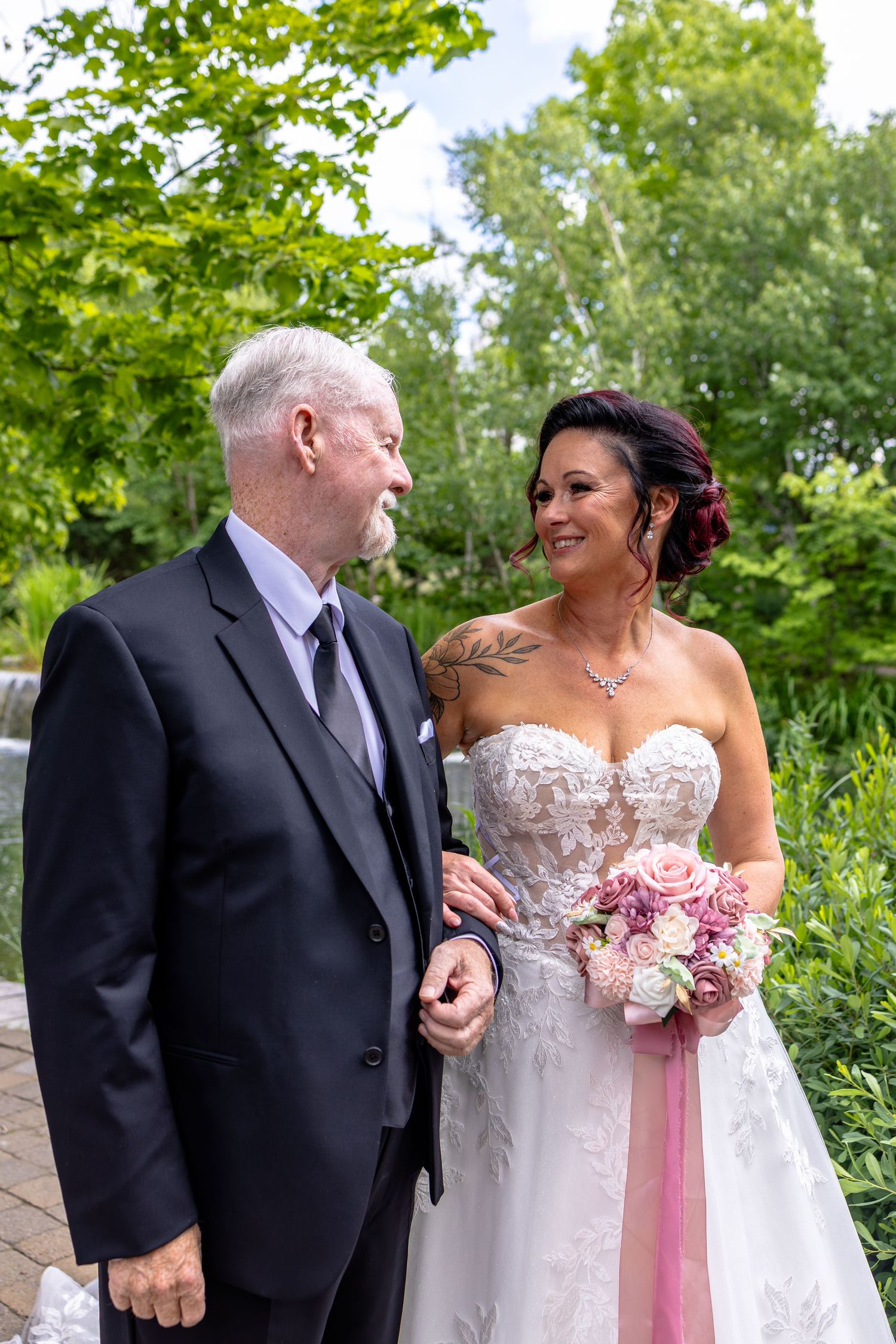 Bride in white lace gown holding pink bouquet stands with groom in dark suit outdoors at wedding ceremony.