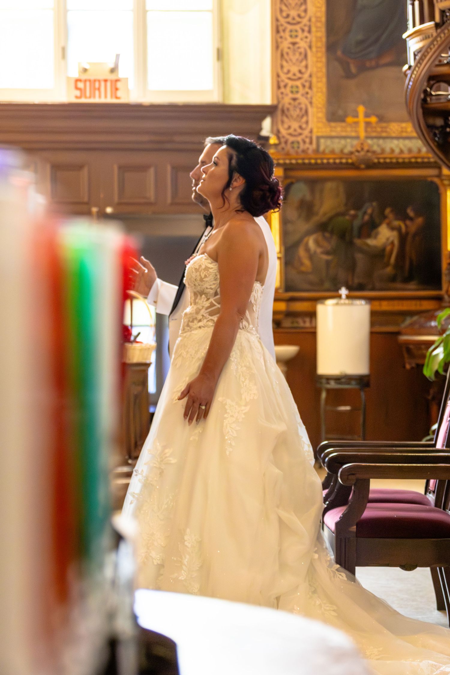 Bride in elegant white gown standing in ornate church, looking upward with joy during wedding ceremony.