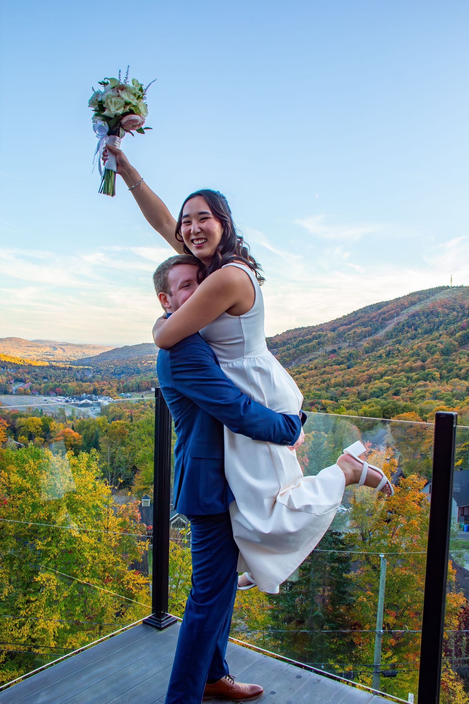 Groom lifts smiling bride who raises her bouquet triumphantly on a balcony with autumn mountain backdrop.