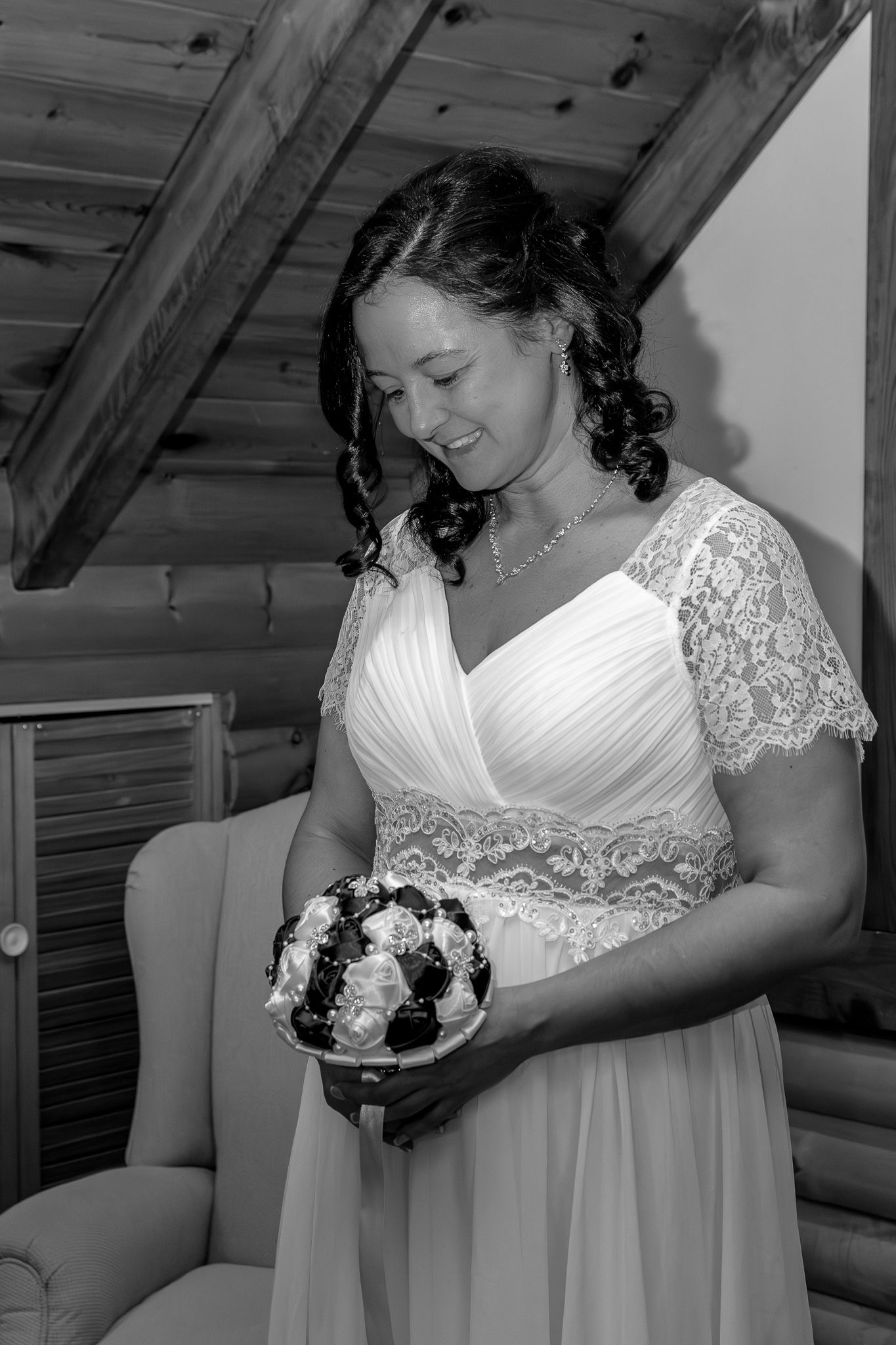 Black and white portrait of smiling bride in lace wedding gown holding bouquet in log cabin.