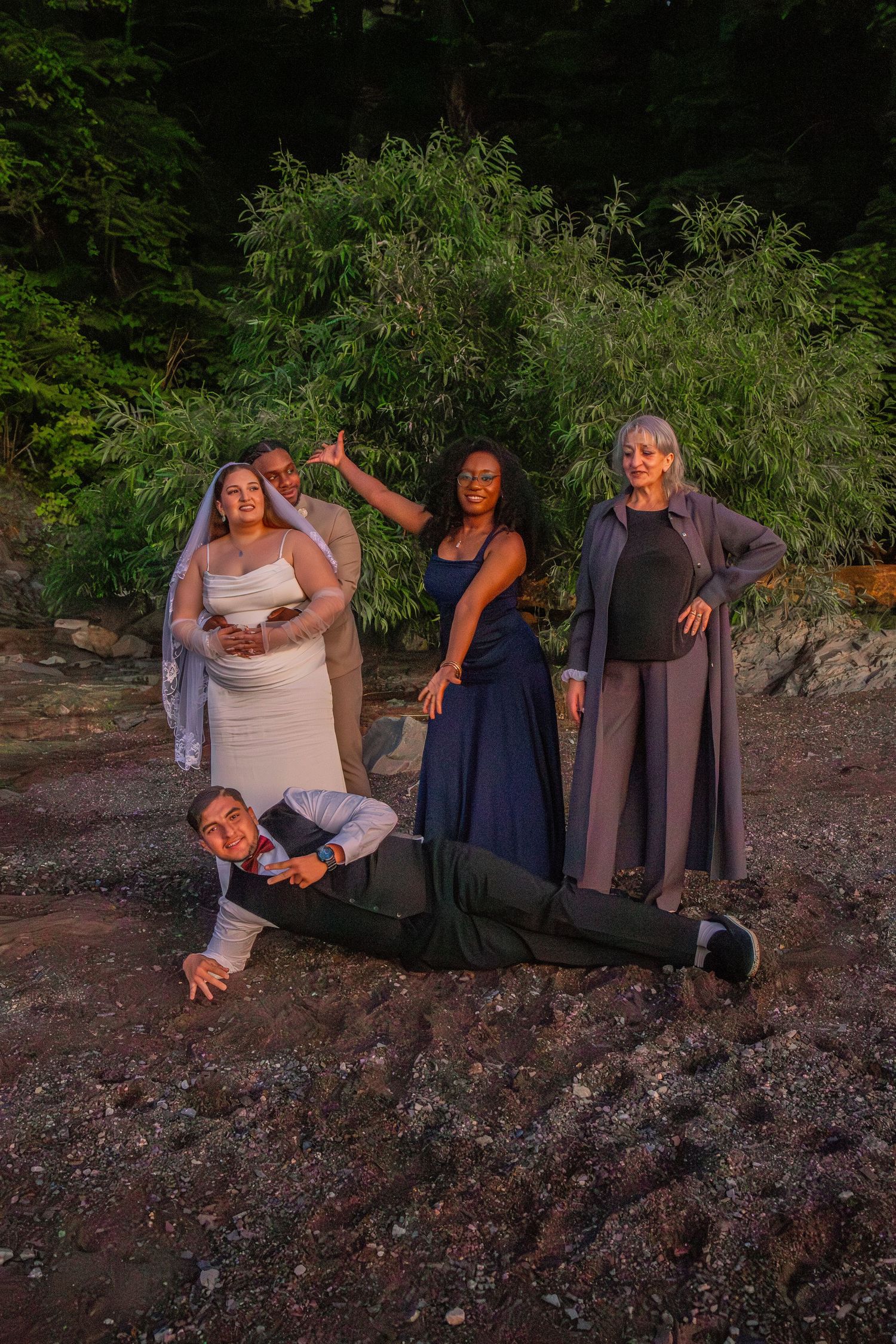 Wedding party posing outdoors, bride in white veil, groom lying on ground, bridesmaids in navy and gray, forest backdrop.