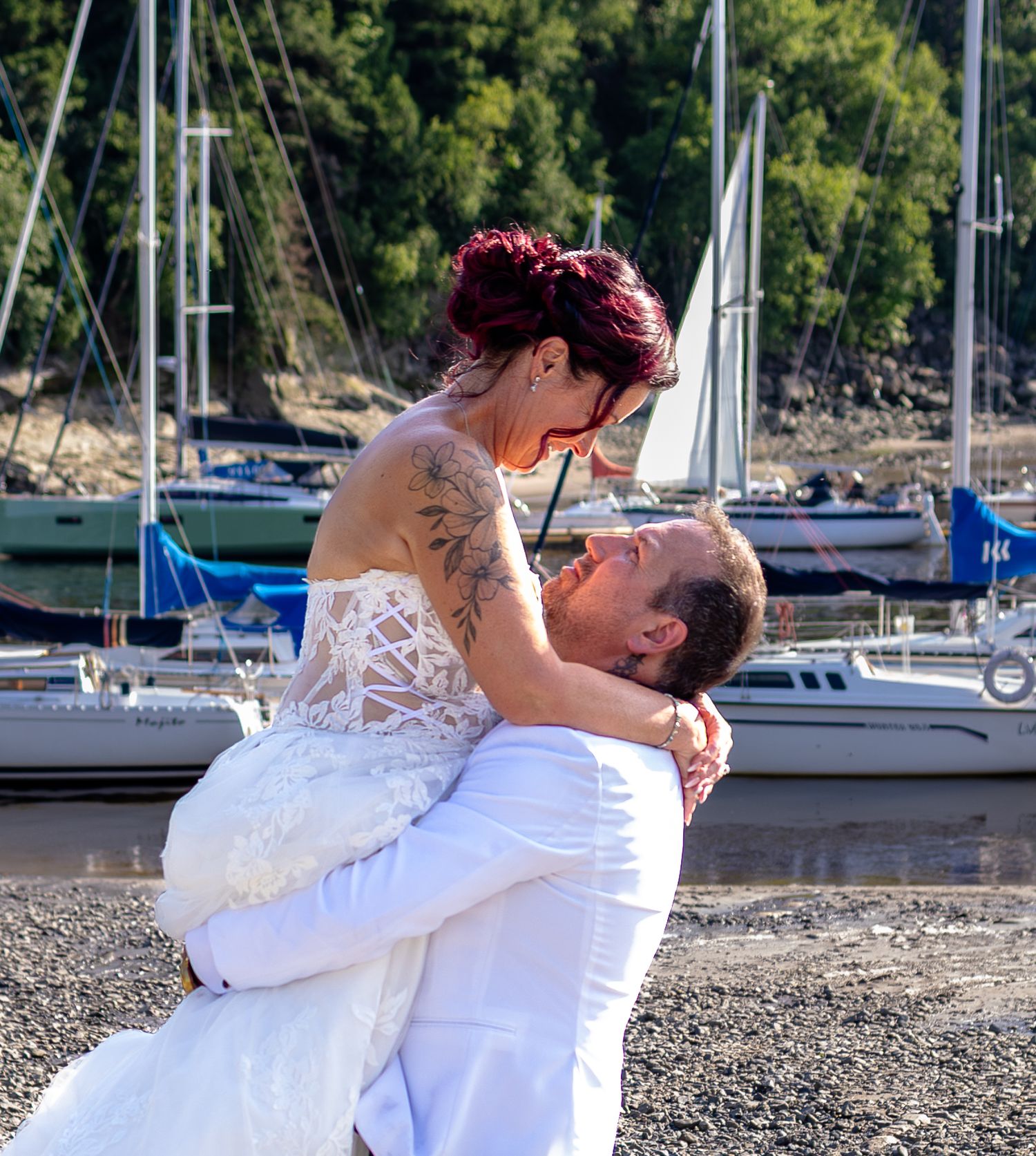 Groom in white lifts bride in lace wedding dress near sailboats on a rocky shoreline.