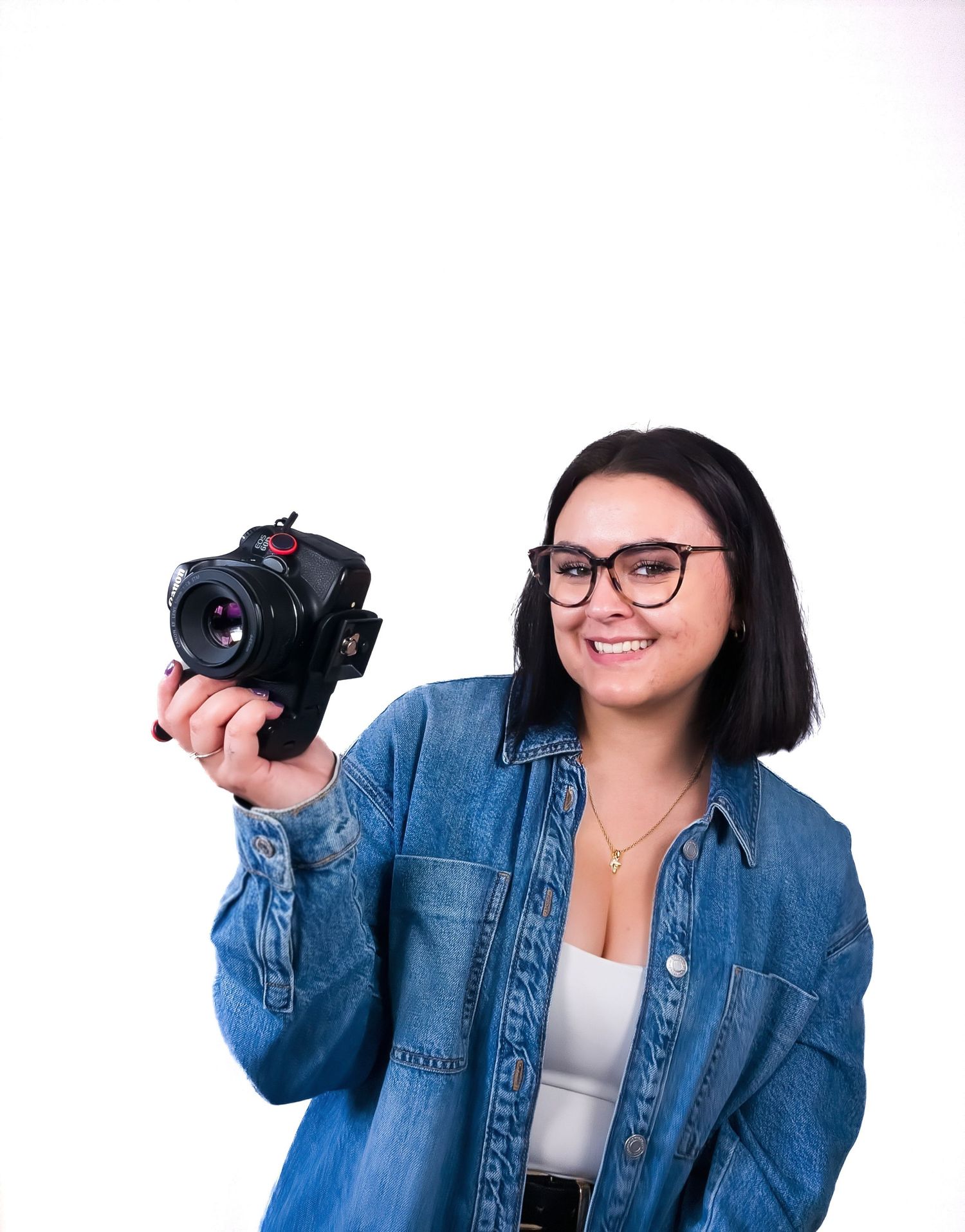 Smiling woman with glasses in denim jacket proudly holding a professional camera against a white background.