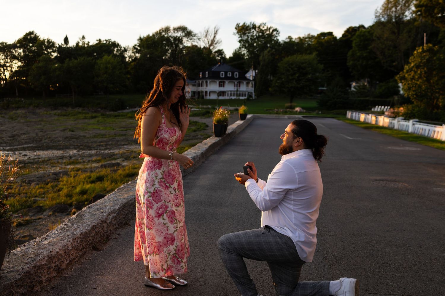 Man proposes on one knee to woman in floral dress on a scenic road at sunset near a historic house.