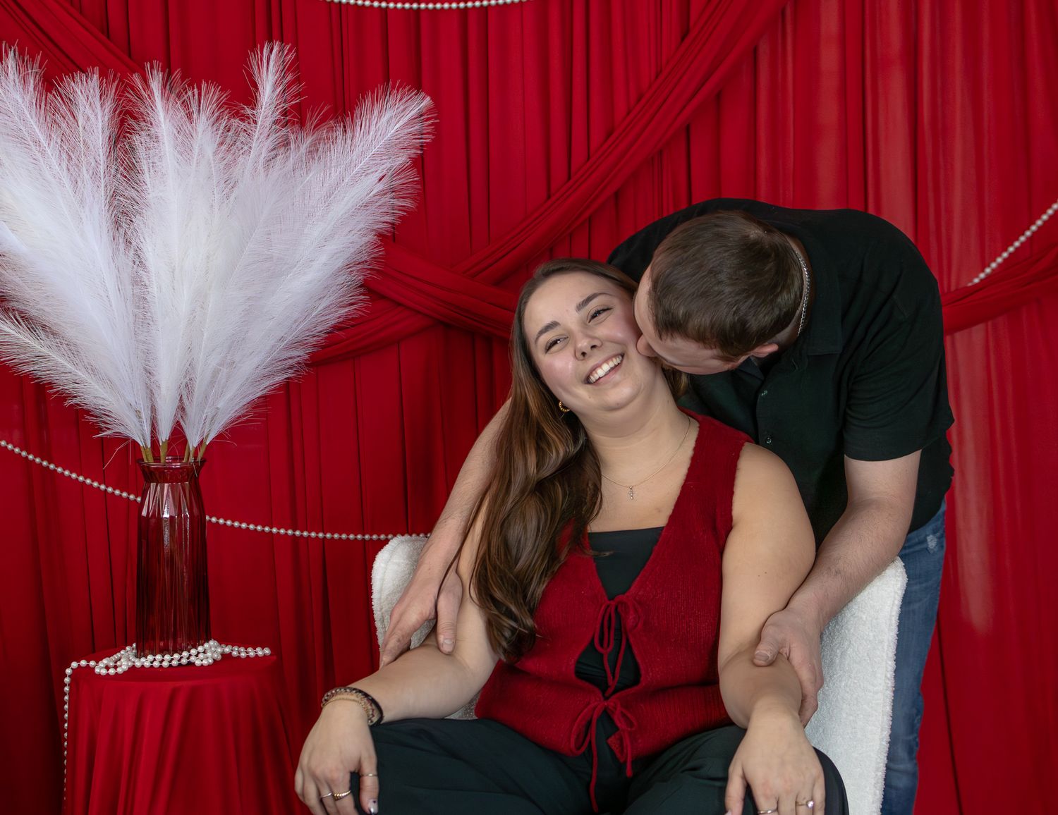 Man kissing smiling woman on cheek as she sits in white chair before red curtain backdrop.