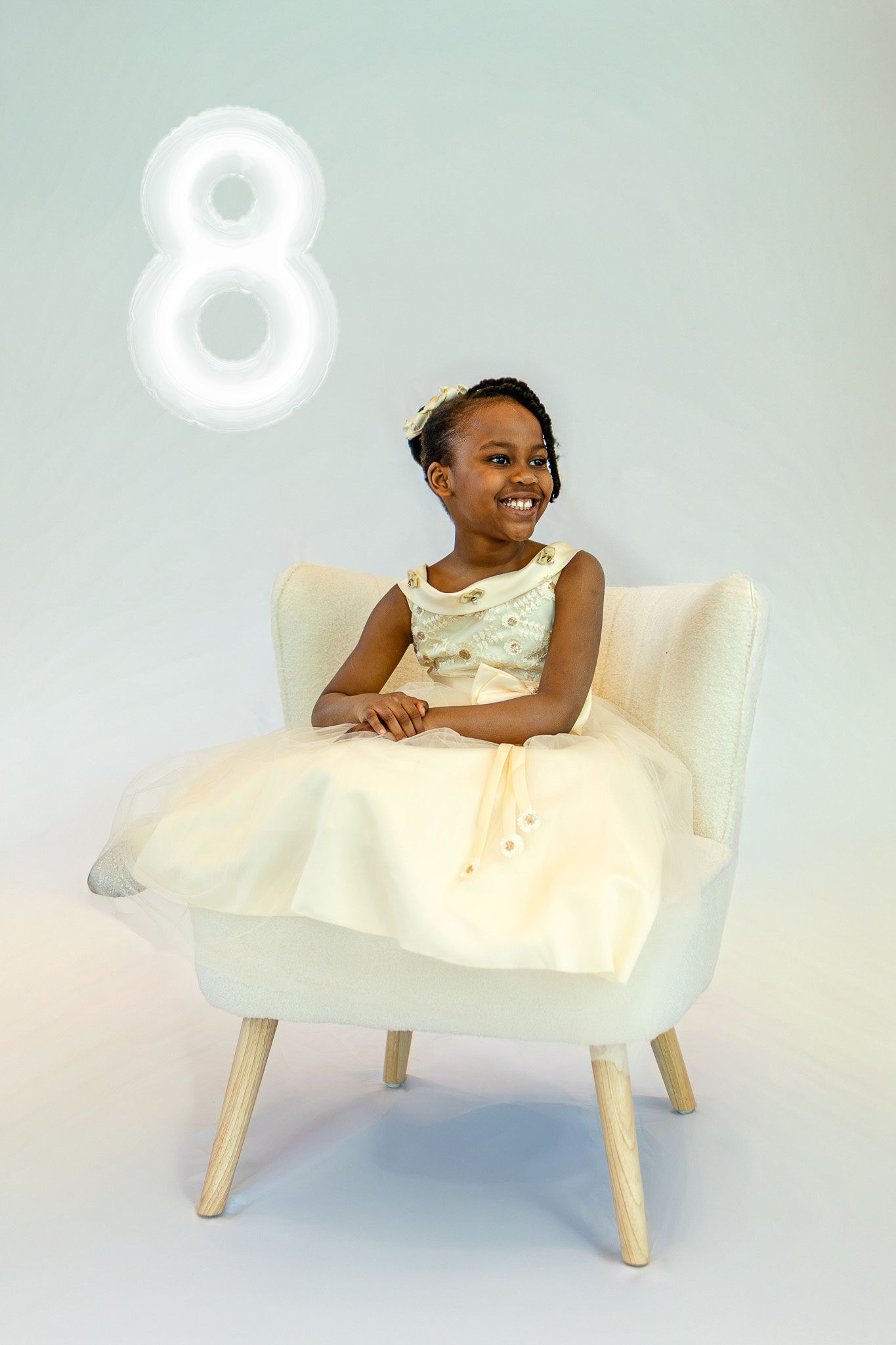 Smiling young Black girl in cream dress sitting in white chair with glowing number 8 above her.